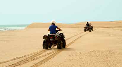 To personer kører på firehjulstrækkere over en sandstrand med havet i baggrunden. (AI-genereret tekst)