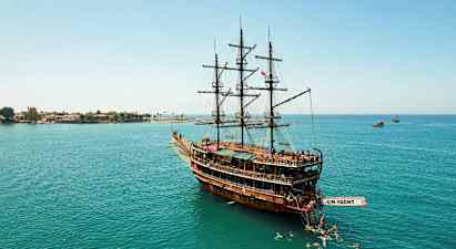 Et stort træskib med sejl, der ligger stille på vandet, omgivet af svømmere, og kystlinjen kan ses i baggrunden. (AI-genereret tekst)