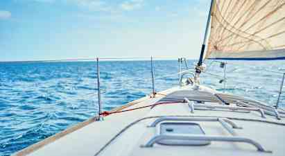 Nærbillede af fordækket på en sejlbåd, med et hvidt sejl og blåt hav i baggrunden. (AI-genereret tekst)