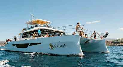 Personer på en katamaran i solrigt vejr, omgivet af vand og en kystlinje i baggrunden. (AI-genereret tekst)