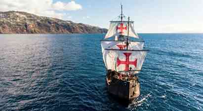 Et sejlskib med store hvide sejl dekoreret med røde kors sejler over et blåt hav nær en kuperet kyst. (AI-genereret tekst)