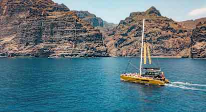 En katamaran sejler tæt på høje klipper, med klart blåt vand i forgrunden. (AI-genereret tekst)