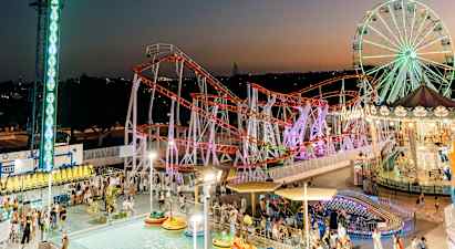 Et aftensbillede af en livlig forlystelsespark med et stort pariserhjul, en farverig rutsjebane og en pool med radiobiler. (AI-genereret tekst)