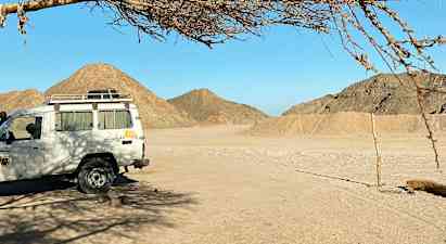 En hvid bil holder parkeret under et træ i et ørkenområde med bjerge i baggrunden. (AI-genereret tekst)