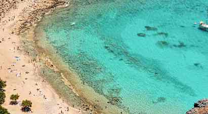 Strand med gyldent sand og klart turkis vand. Mange mennesker nyder solen og svømmer. En sejlbåd ligger for anker længere ude. (AI-genereret tekst)