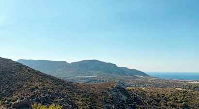 En udsigt over et bjergrigt landskab med varieret vegetation og havet i baggrunden under en klar blå himmel. (AI-genereret tekst)