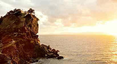 En klippe ved kysten med en bygning og træer på toppen. Havet ses under en solnedgang. (AI-genereret tekst)