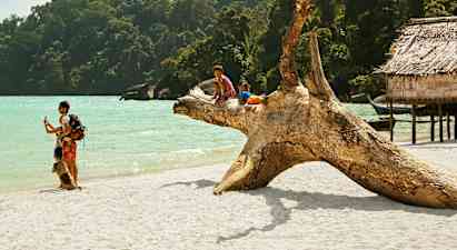 To børn sidder på en stor træstub på en sandstrand ved havet, omgivet af frodig skov og træbygninger. (AI-genereret tekst)