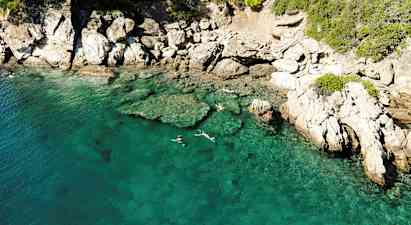 Klart turkisblåt vand ved en klippefuld kyst, hvor personer snorkler nær overfladen. (AI-genereret tekst)