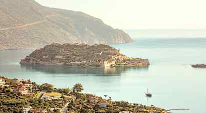 En udsigt over en historisk ø med ruiner og omkringliggende hav, og i forgrunden ses en kystby med bygninger. (AI-genereret tekst)