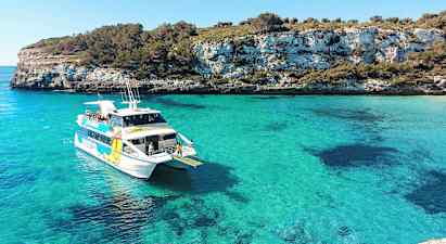 En hvid båd med passagerer i en turkisblå bugt, omgivet af klippeområder med grønt. (AI-genereret tekst)