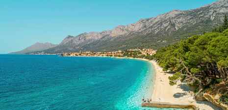 Udsigt over en stenstrand med turkis vand og frodige træer. Bjerge og en landsby ses i baggrunden. (AI-genereret tekst)