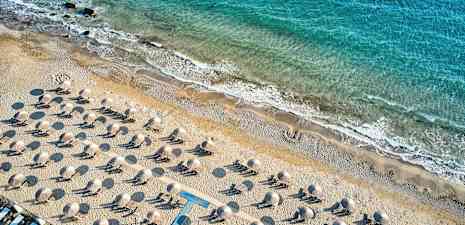 Luftfoto af en sandstrand med liggestole og parasoller, med kystlinjen og blåt hav i baggrunden. (AI-genereret tekst)