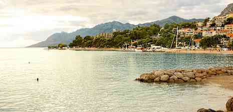 Strand med klart vand, stenbrygge og omkringliggende bygninger med bjerge i baggrunden. (AI-genereret tekst)