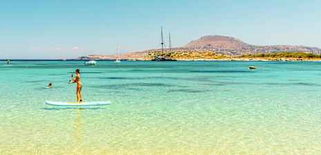 Person, der står på et paddleboard i klart blåt vand med en bjergfyldt kystlinje i baggrunden. (AI-genereret tekst)