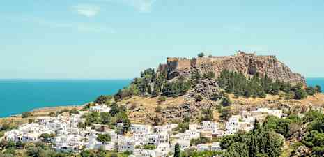 Udsigt over en bakke med en historisk fæstning omgivet af grønne træer og hvide bygninger nær havet. (AI-genereret tekst)