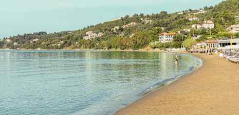 En rolig strand ved kysten med klart blåt vand, grønne træer og huse på en skråning. (AI-genereret tekst)