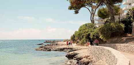 Stenstrand med snoet gangsti tæt på havet, omgivet af træer og bygninger. (AI-genereret tekst)