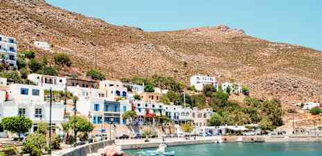 En kystby med hvide og farverige bygninger, små både ved en stenfyldt kystlinje og tørre bjerge i baggrunden. (AI-genereret tekst)