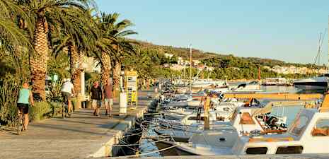 Gåtur ved lystbådehavn med palmer og parkerede både på en solrig dag. (AI-genereret tekst)