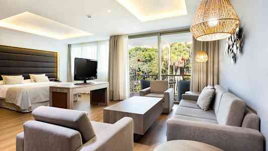 Et hotelværelse, dekorativt møbleret med sofa, lænestole, seng og fjernsyn, samt udsigt til en frodig have. (AI-genereret tekst)