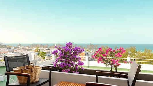 En altan med bord og stole, blomster og havudsigt. (AI-genereret tekst)