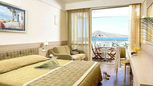 Hotelværelse med seng, siddeområde og udsigt til havet gennem en balkon med stole og bord. (AI-genereret tekst)