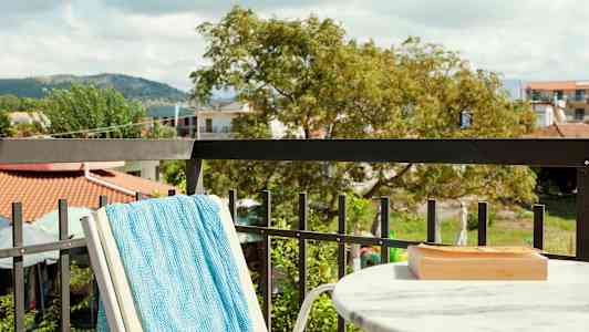 Udsigt fra en altan med et marmorbord, et turkis håndklæde på en stol og grønne omgivelser i baggrunden. (AI-genereret tekst)