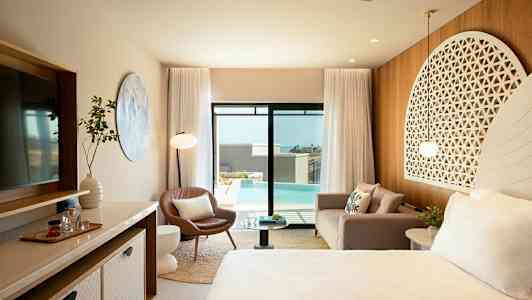 Et elegant hotelværelse med moderne møbler, en sofa, en lænestol, et sengeområde med dekorativ hovedgærde, et stort spejl og glasdøre, der åbner ud til en udendørs swimmingpool. (AI-genereret tekst)