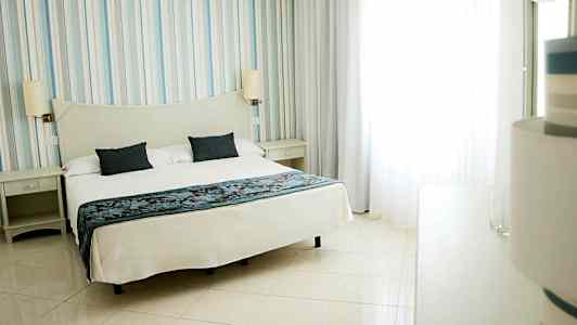 Et hotelværelse med en stor seng dækket med hvidt sengetøj og to mørkeblå puder. (AI-genereret tekst)