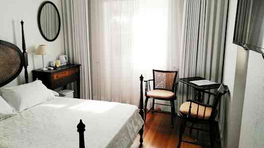 Et hotelværelse med en seng, skrivebord og stol samt et vindue med hvide gardiner. (AI-genereret tekst)