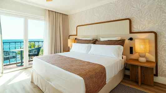 Et hotelværelse med en stor seng, to natborde med lamper og en balkon med udsigt over havet. (AI-genereret tekst)