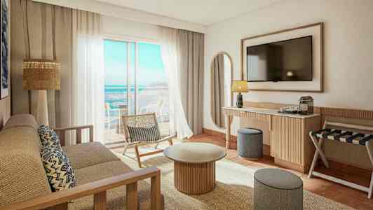 Et lyst og moderne hotelværelse med udsigt over havet. Der er en sofa, et sofabord, en stol og et fjernsyn på væggen. (AI-genereret tekst)