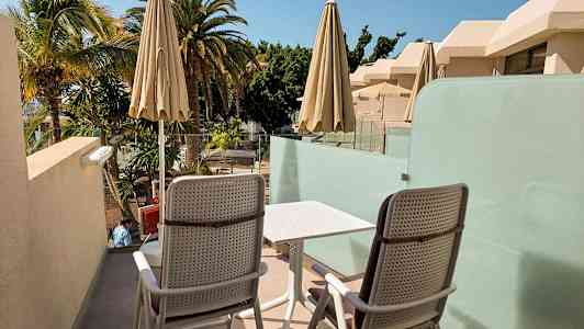 To stole og et bord på en terrasse med udsigt over palmer og bygninger. (AI-genereret tekst)