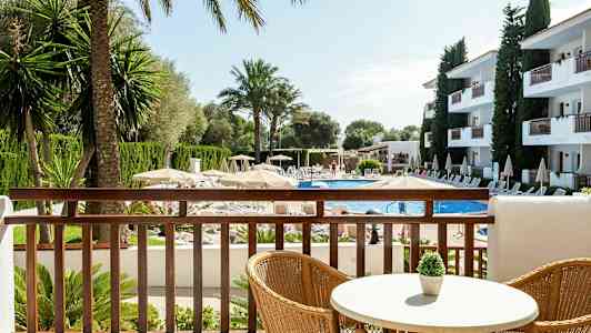 Udsigt fra en balkon over en pool omgivet af liggestole, parasoller og bygninger. (AI-genereret tekst)