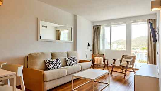 En stue med en beige sofa, to stole og et hvidt sofabord. Der er et vindue med udsigt til grønne omgivelser. (AI-genereret tekst)