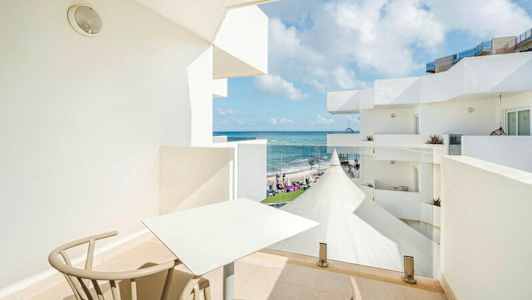 En balkon med bord og stole, med udsigt til havet og hvide bygninger. (AI-genereret tekst)