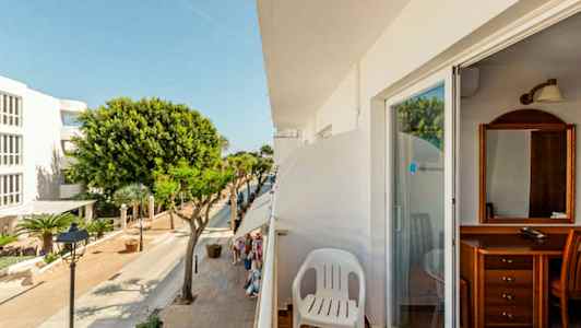 Udsigt fra en altan med en hvid plaststol. Gaden nedenfor har træer og bygninger. (AI-genereret tekst)