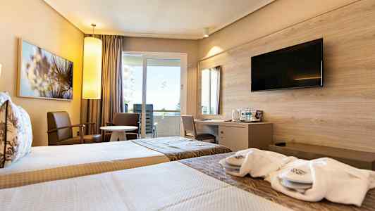 Et hotelværelse med to enkeltsenge, siddepladser ved et rundt bord samt en skrivebordsafdeling med en vægmonteret TV over. (AI-genereret tekst)