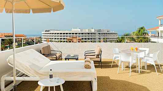Udendørs terrasse med parasol, liggestole og siddeområde. Havet og en bygning kan ses i baggrunden. (AI-genereret tekst)