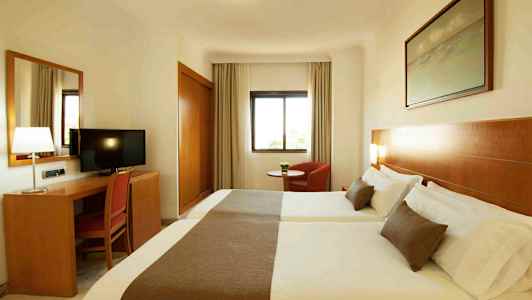 Et hotelværelse med to senge, et skrivebord med en stol og et fjernsyn, samt et vindue og en lænestol. (AI-genereret tekst)