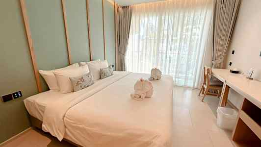 Billede af et moderne hotelværelse med en stor redt seng, hvide sengeklæder og dekorative puder samt håndklæder formet som svaner. Værelset har vinduer med lyse gardiner og et skrivebord ved siden af. (AI-genereret tekst)