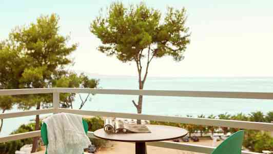 En altan med grønne stole, et bord og udsigt over træerne og havet. (AI-genereret tekst)