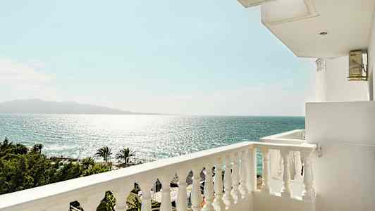 Udsigt fra en balkon over havet og en klar himmel, med palmer og trækroner nederst. (AI-genereret tekst)