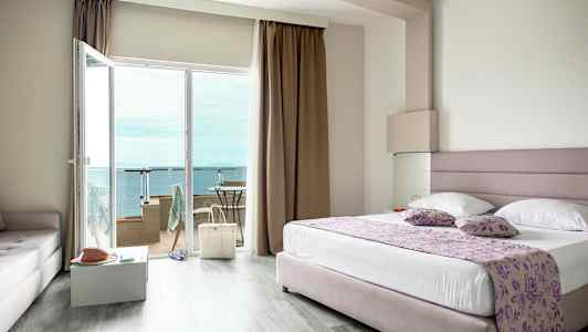 Et hotelværelse med en stor seng, glasdøre til en balkon og udsigt over havet. (AI-genereret tekst)