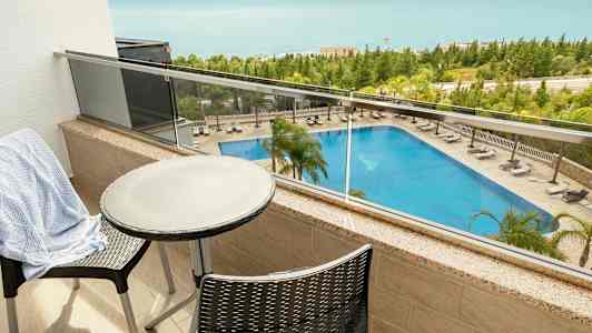 Udsigt fra en balkon med bord og stole, der viser en pool og havet i baggrunden. (AI-genereret tekst)