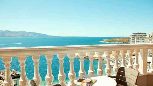 Udsigt fra en balkon med et hvidt rækværk over havet, et bord og en stol. (AI-genereret tekst)