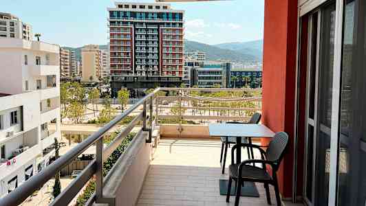 En altan med bord og stole med udsigt over omkringliggende bygninger og bjerge i det fjerne. (AI-genereret tekst)