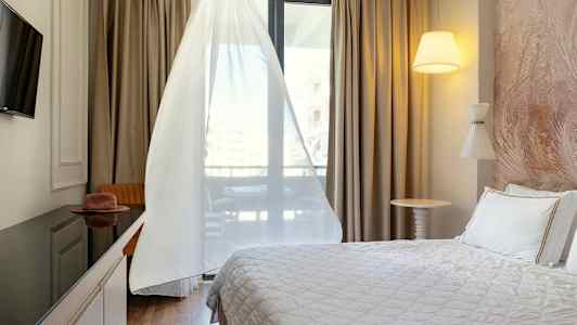 Et hotelværelse med en seng, TV, bord og stol samt et vindue med hvide gardiner. (AI-genereret tekst)