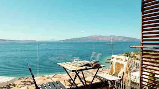 Udsigt fra en altan mod havet og en strand med parasoller. På altanen står et lille bord og en stol. (AI-genereret tekst)